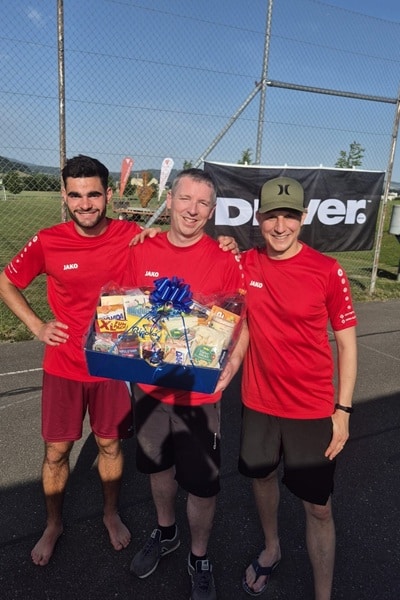 Drei Personen in roten Sportshirts halten einen Geschenkkorb und lachen in die Kamera